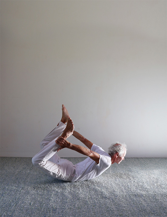Yogaübender in weißer Kleidung zeigt Dhanurasana, den Bogen, mit angehobenen Beinen und einer Rückwärtsbeuge der Wirbelsäule.